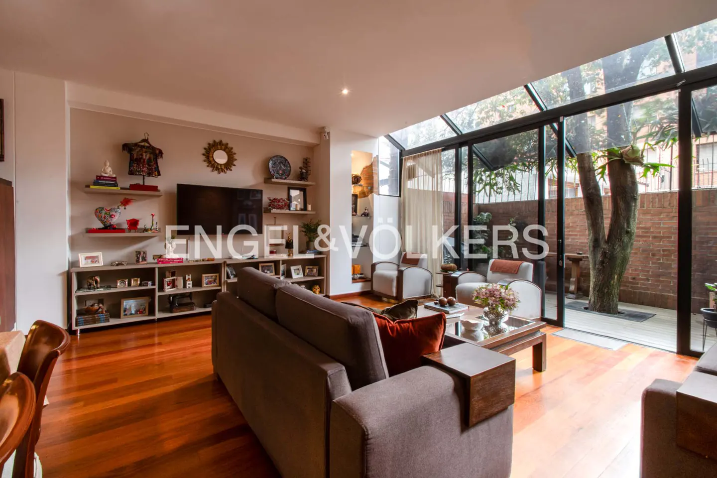 Living room with hardwood floors, a brown sofa, and a glass-enclosed patio with a tree. Shelves and a TV are on the wall.