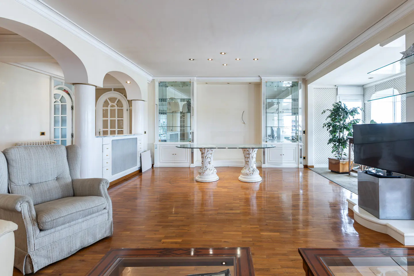 Bright living room with hardwood floors, a gray armchair, glass table, and built-in white cabinets. Arched doorways and a large TV add to the room's elegance.