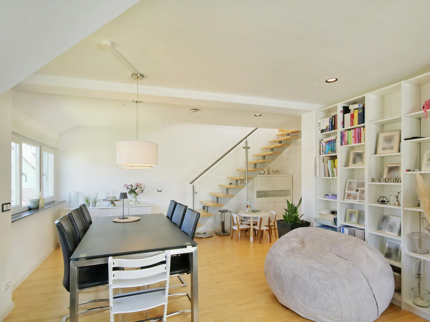 Bright, modern living space with wood floors, a dining table with black chairs, a staircase, and a large white bookshelf.