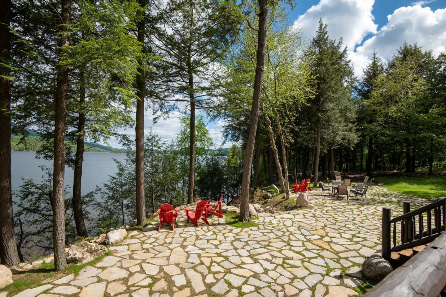 Outdoor patio with red Adirondack chairs and a table set overlooking a lake surrounded by tall green trees.
