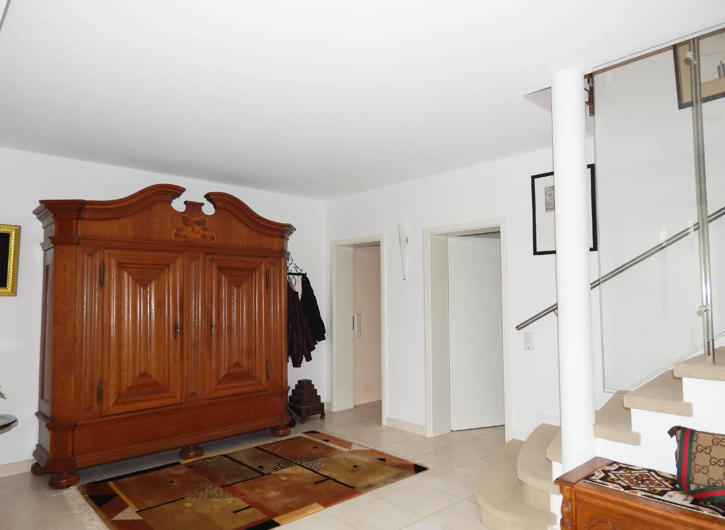 Bright foyer with a large, ornate wooden armoire, a patterned rug, and a staircase with glass railings. White walls and doors add to the airy feel.