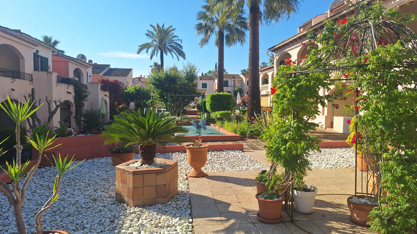 Courtyard view of a Mediterranean-style complex with palm trees, potted plants, and white gravel. Buildings are peach-colored with arched balconies.