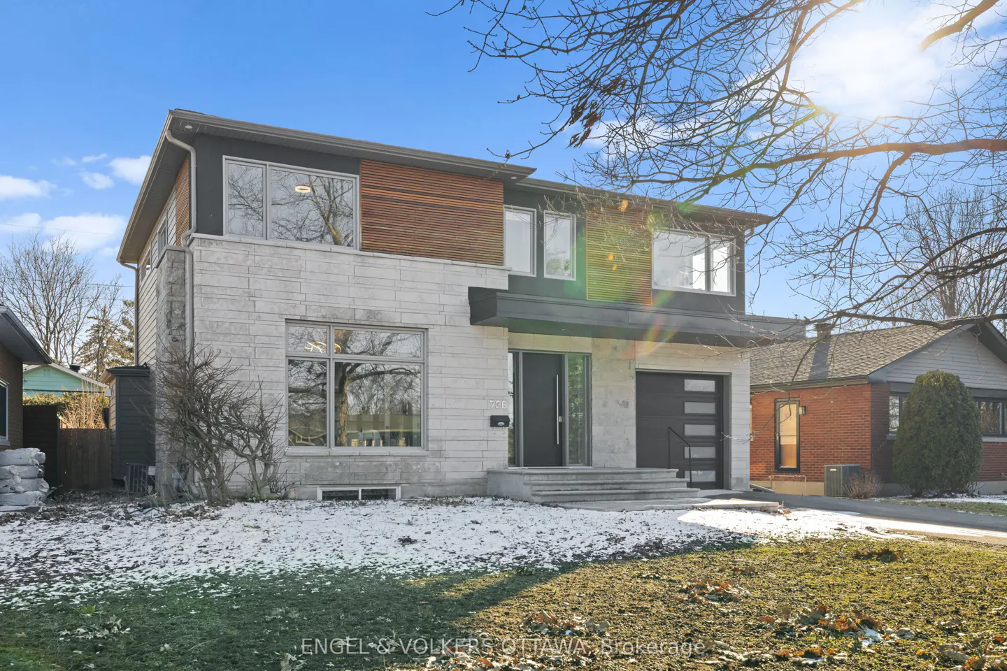 Two-story house with stone and wood exterior, black door and garage, and snowy lawn under a bright blue sky.