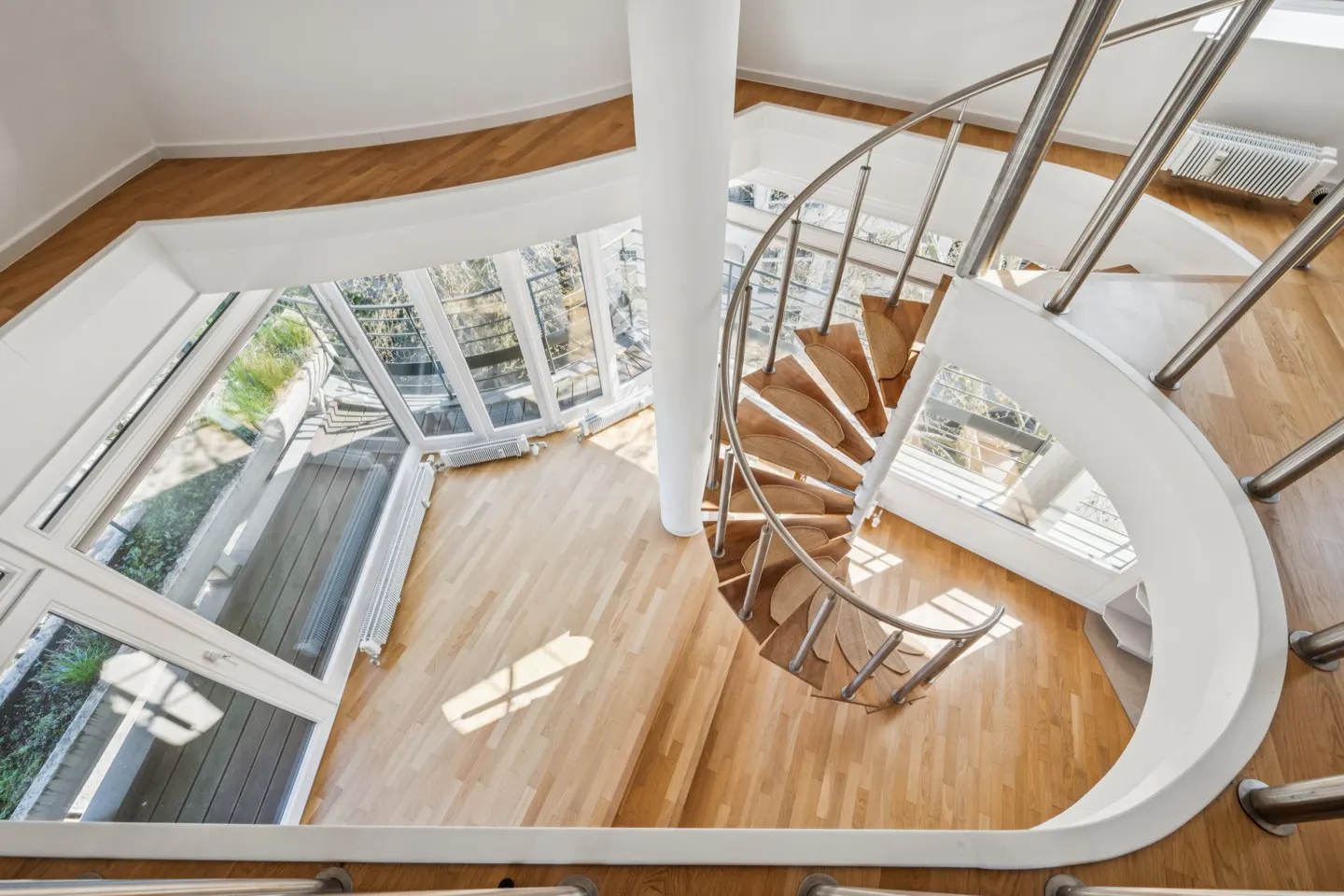 Overhead shot of a modern home's spiral staircase with wood steps and stainless steel railings, next to large windows.