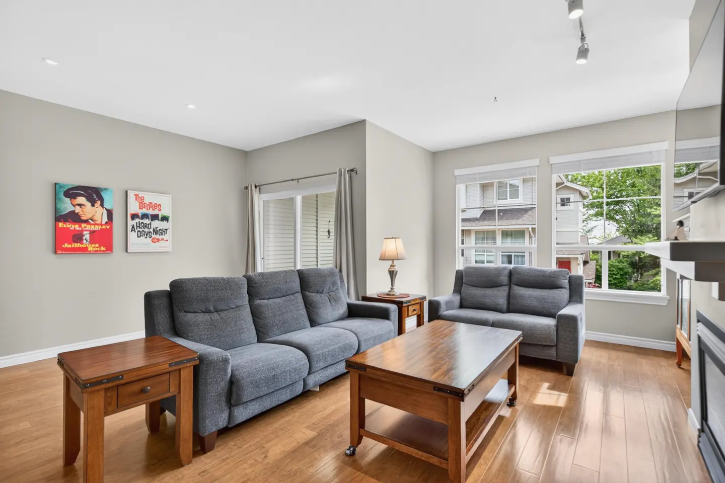 Living room with gray sofas, wood floors, and a coffee table. Elvis and Beatles posters hang on the wall. Windows show a view of houses outside.