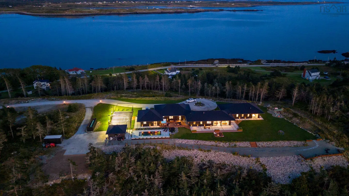 Aerial view of a large, modern home with a dark roof, surrounded by trees and overlooking a blue bay at dusk. A tennis court and pool are visible.