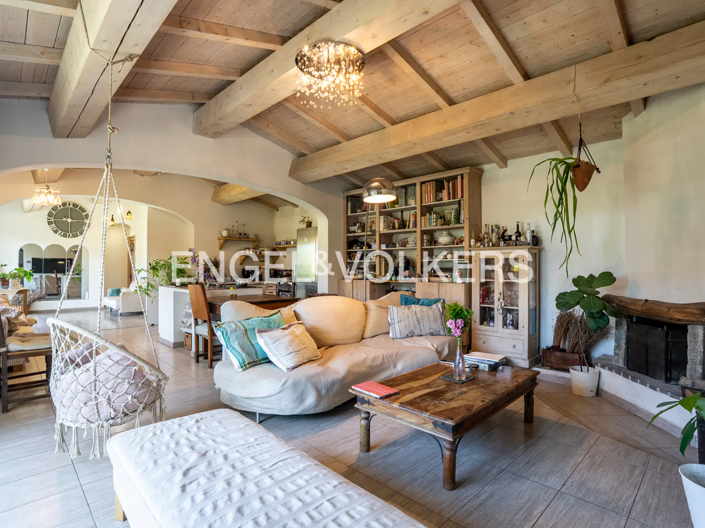 Bright living room with exposed beams, a beige sofa, a wooden coffee table, and a hanging macrame chair. Engel & Volkers logo visible.