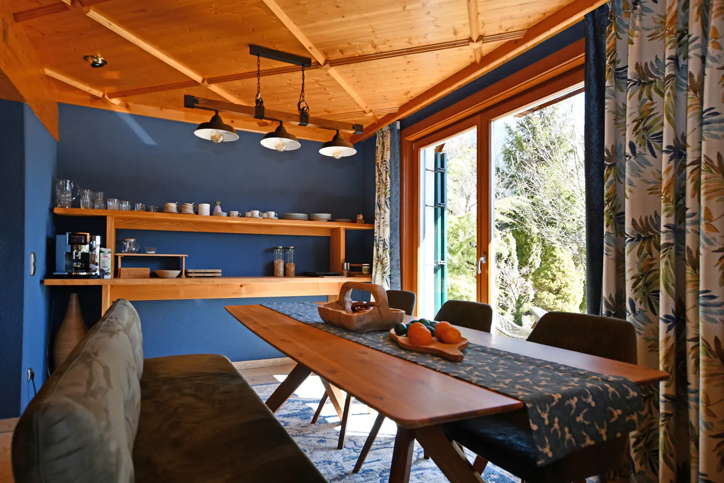 A dining room with a wooden table, bench, and chairs. A blue wall with wooden shelves and a window with floral curtains.