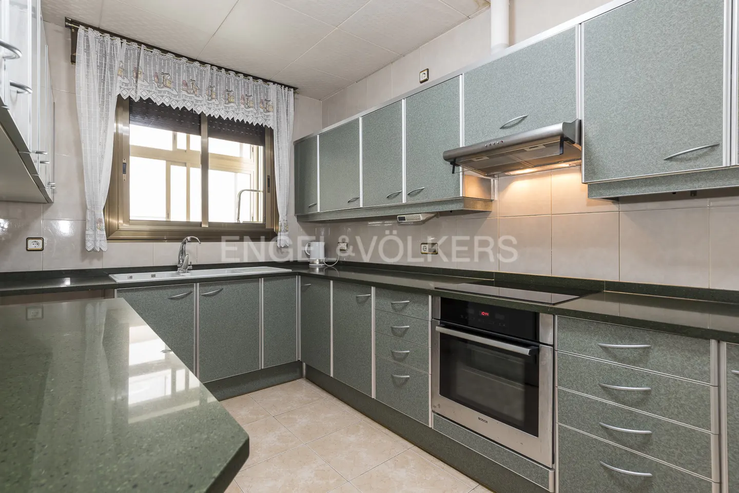 A kitchen with gray cabinets, dark countertops, and a window with white curtains. An oven is built into the cabinets.