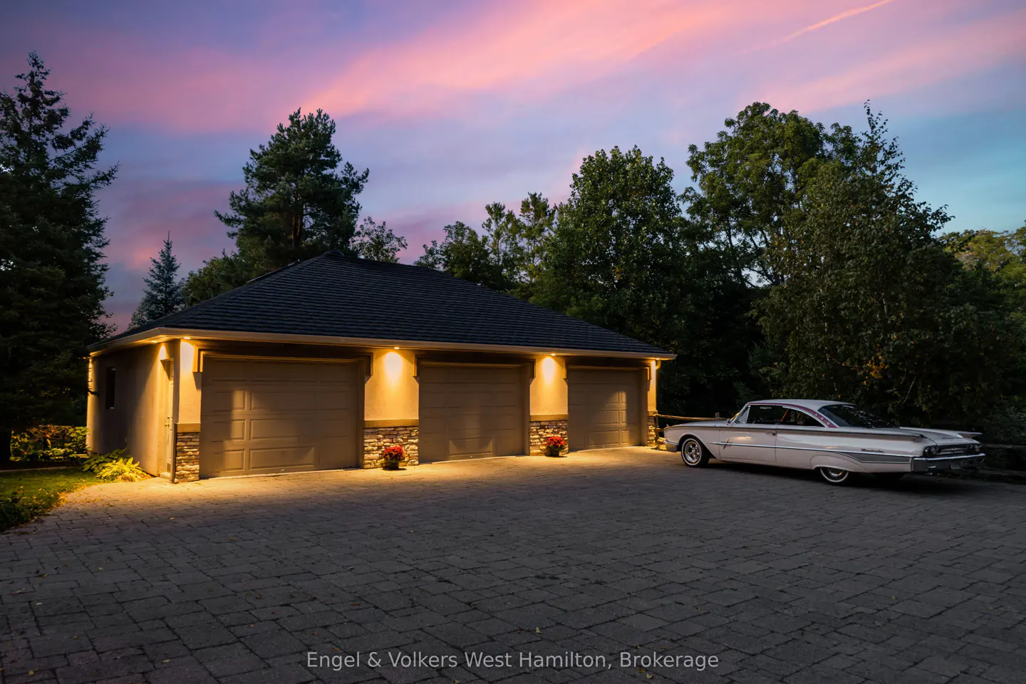 Three-car garage with a vintage white car parked outside on a brick driveway at dusk.