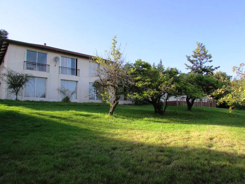 Two-story beige building with balconies, large windows, and a satellite dish, surrounded by green grass and trees under a blue sky.