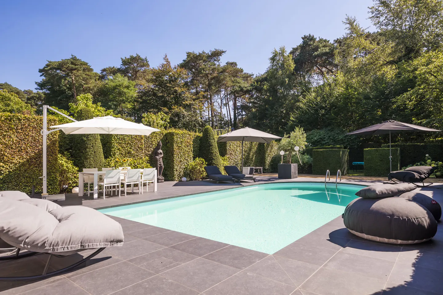 Outdoor pool area with gray tile, surrounded by green trees and hedges. White table and chairs under a white umbrella. Lounge chairs and beanbag chairs are nearby.