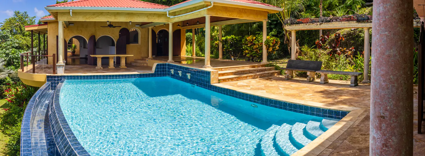A yellow villa with a red roof overlooks a blue-tiled pool with steps. A stone bench sits on a patio with columns and lush greenery.