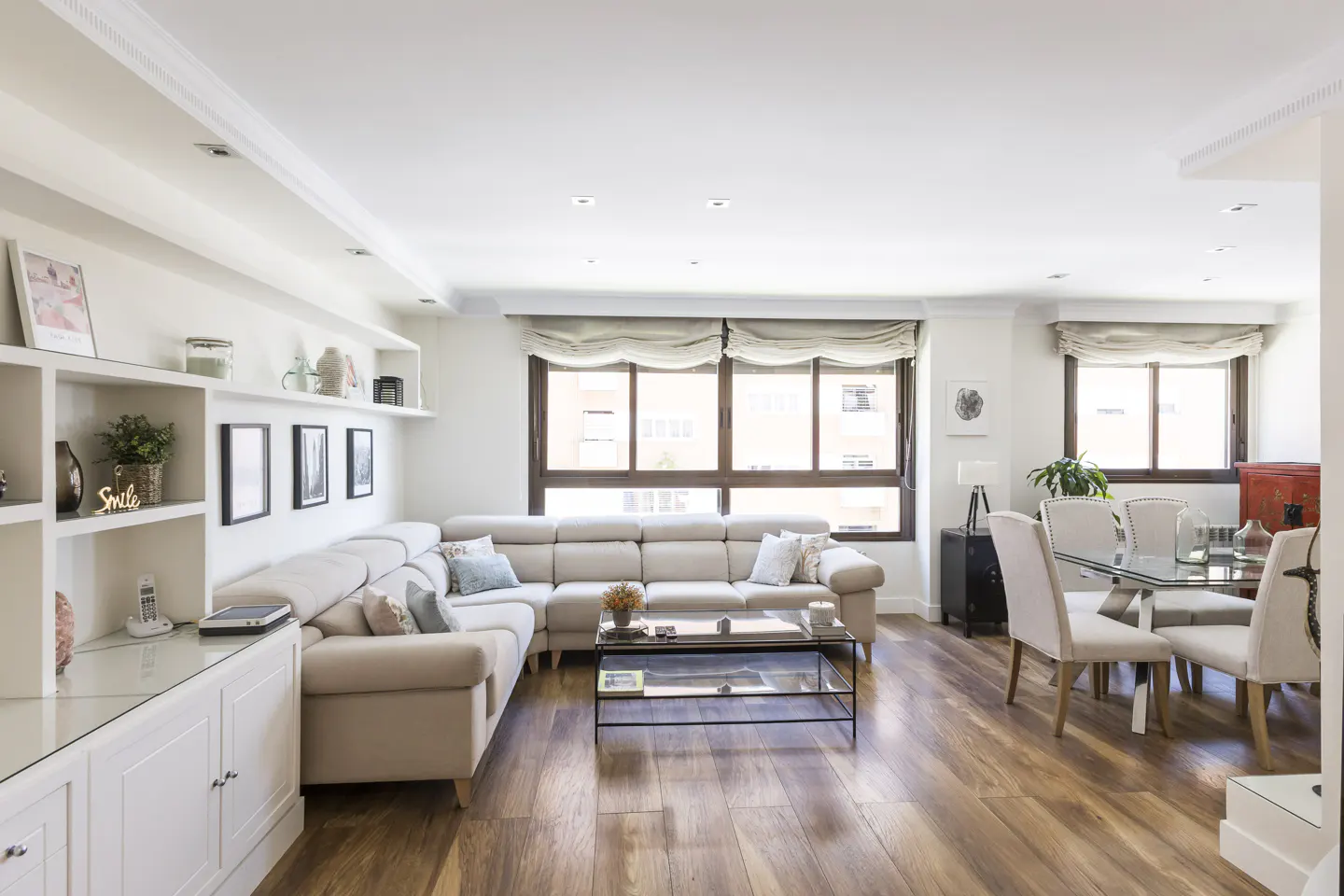 Bright living room with hardwood floors, a beige sectional sofa, and a glass dining table with white chairs. White built-in shelves and large windows add to the airy feel.