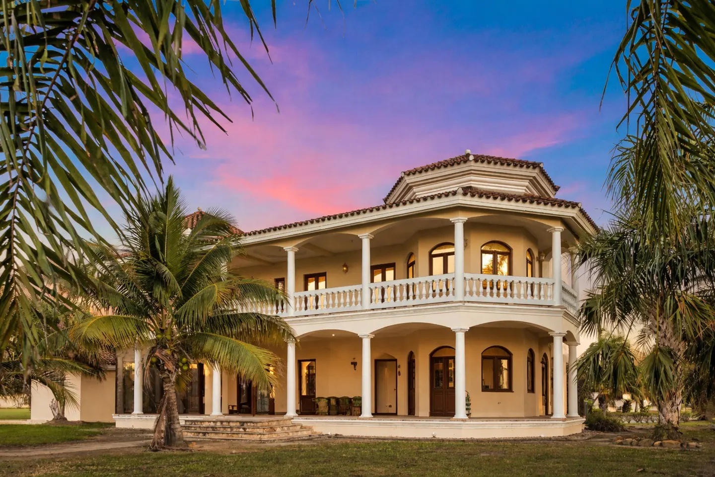 Two-story beige house with white columns and balconies, framed by palm trees against a pink and blue sunset.