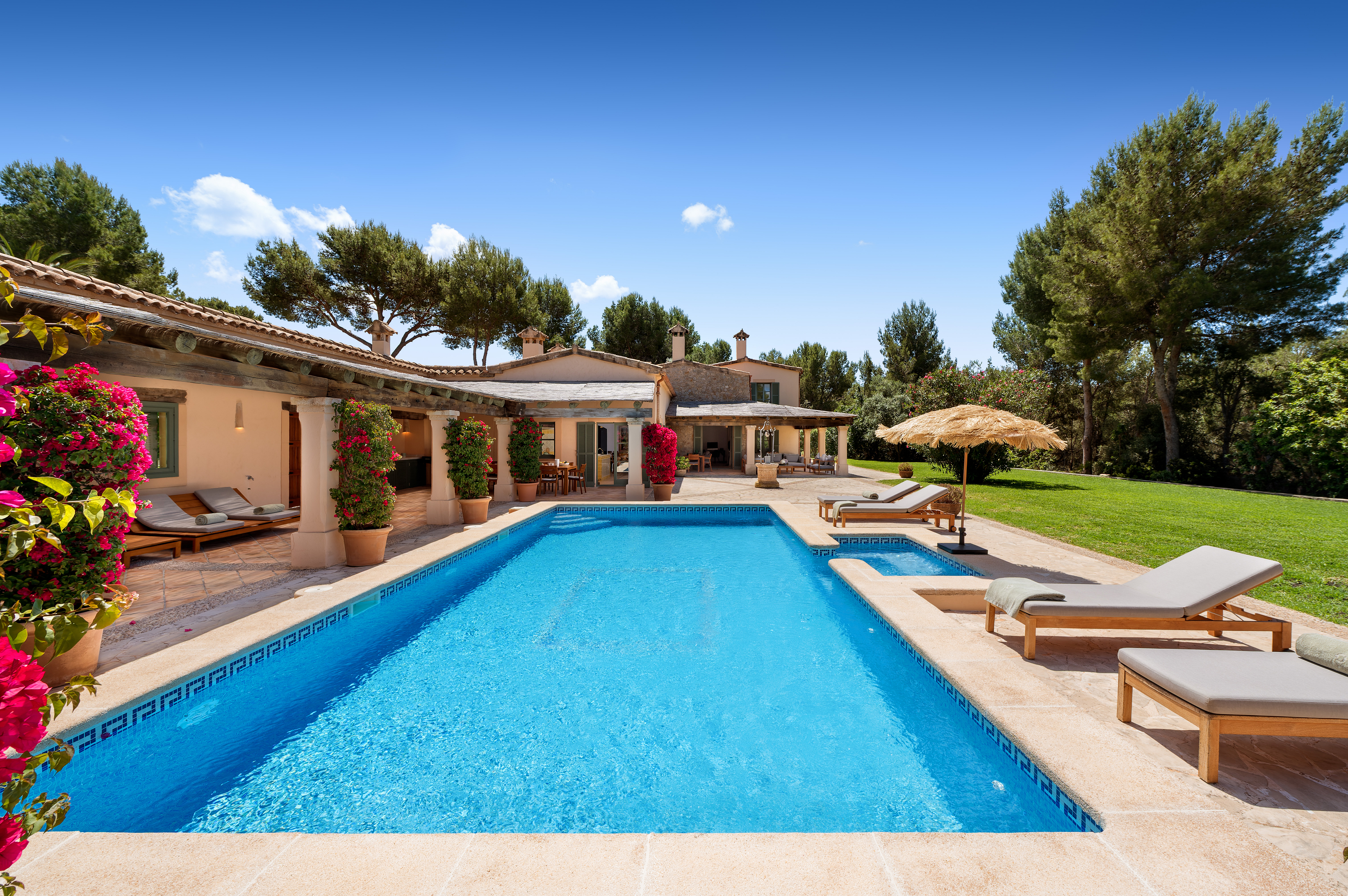A bright blue swimming pool with a stone patio, lounge chairs, and a villa with pink bougainvillea under a clear blue sky.
