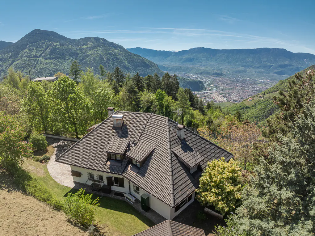 Aerial view of a white house with a brown tiled roof, surrounded by green trees and mountains in the background.