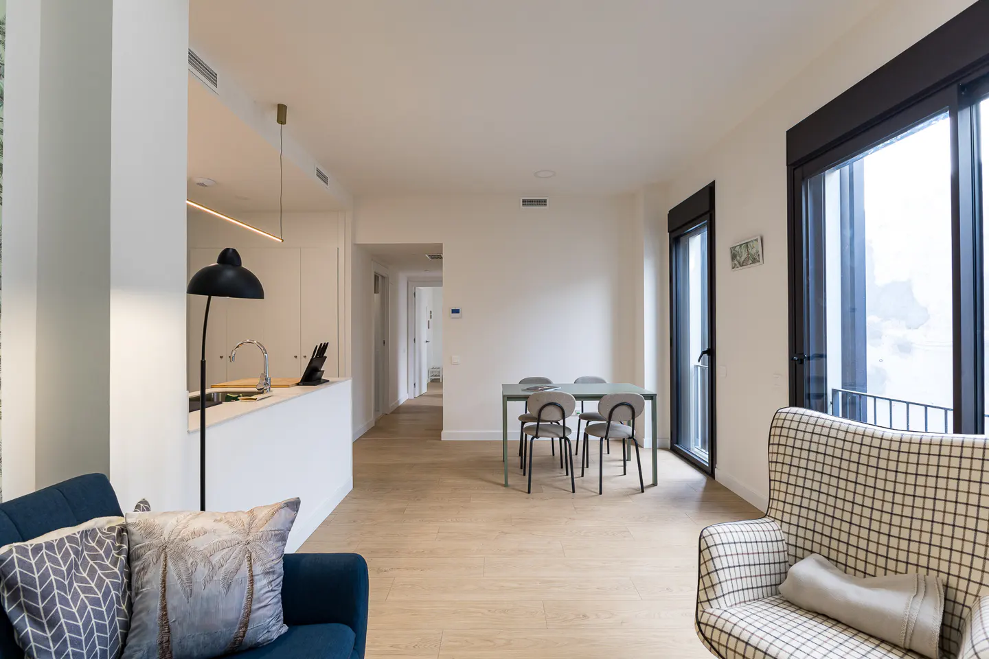 Bright, modern apartment interior with a blue sofa, plaid armchair, dining table, and kitchen counter. Light wood floors and large windows.