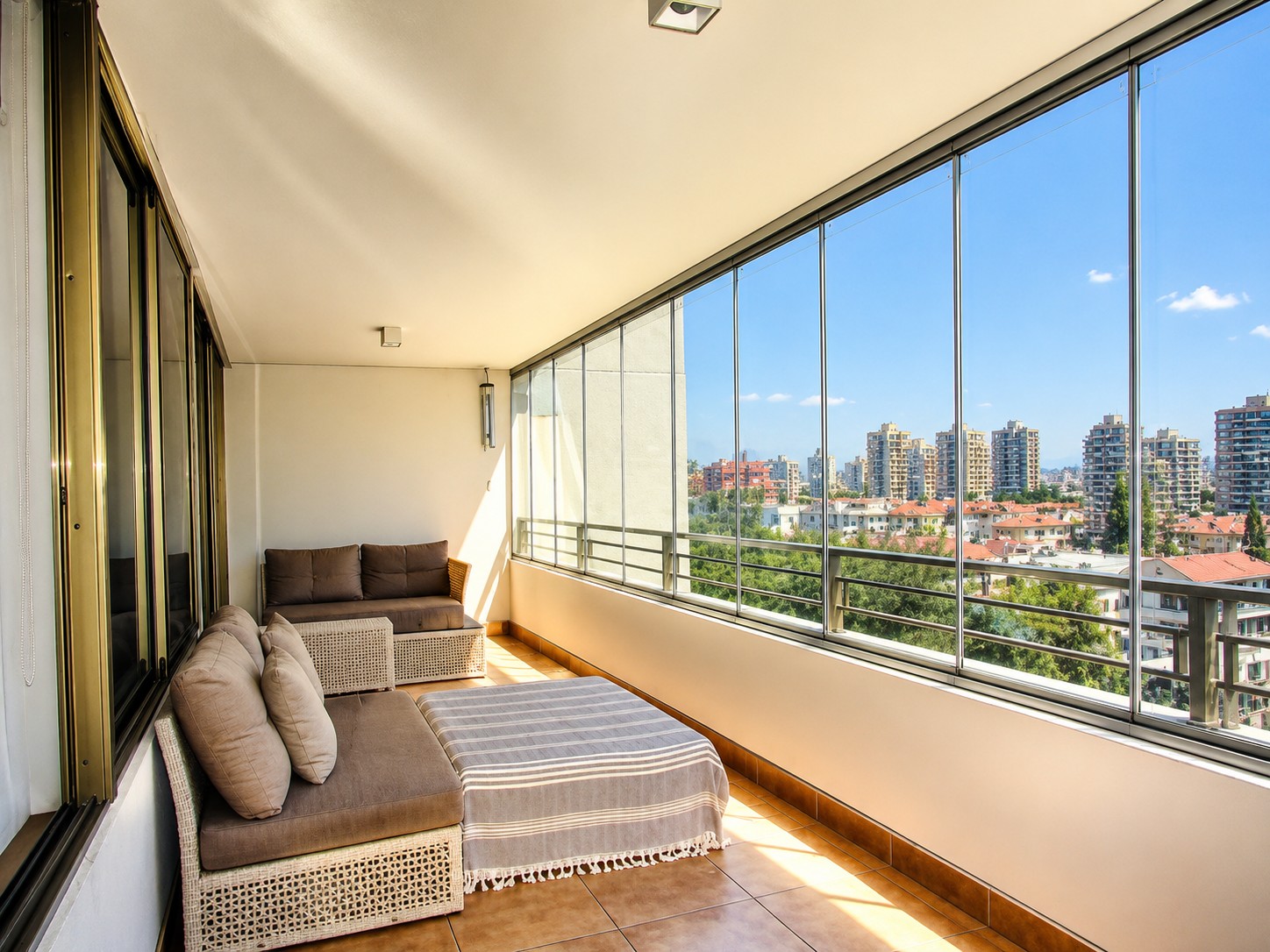 Enclosed balcony with wicker furniture, brown cushions, and a striped throw, overlooking a sunny city skyline.