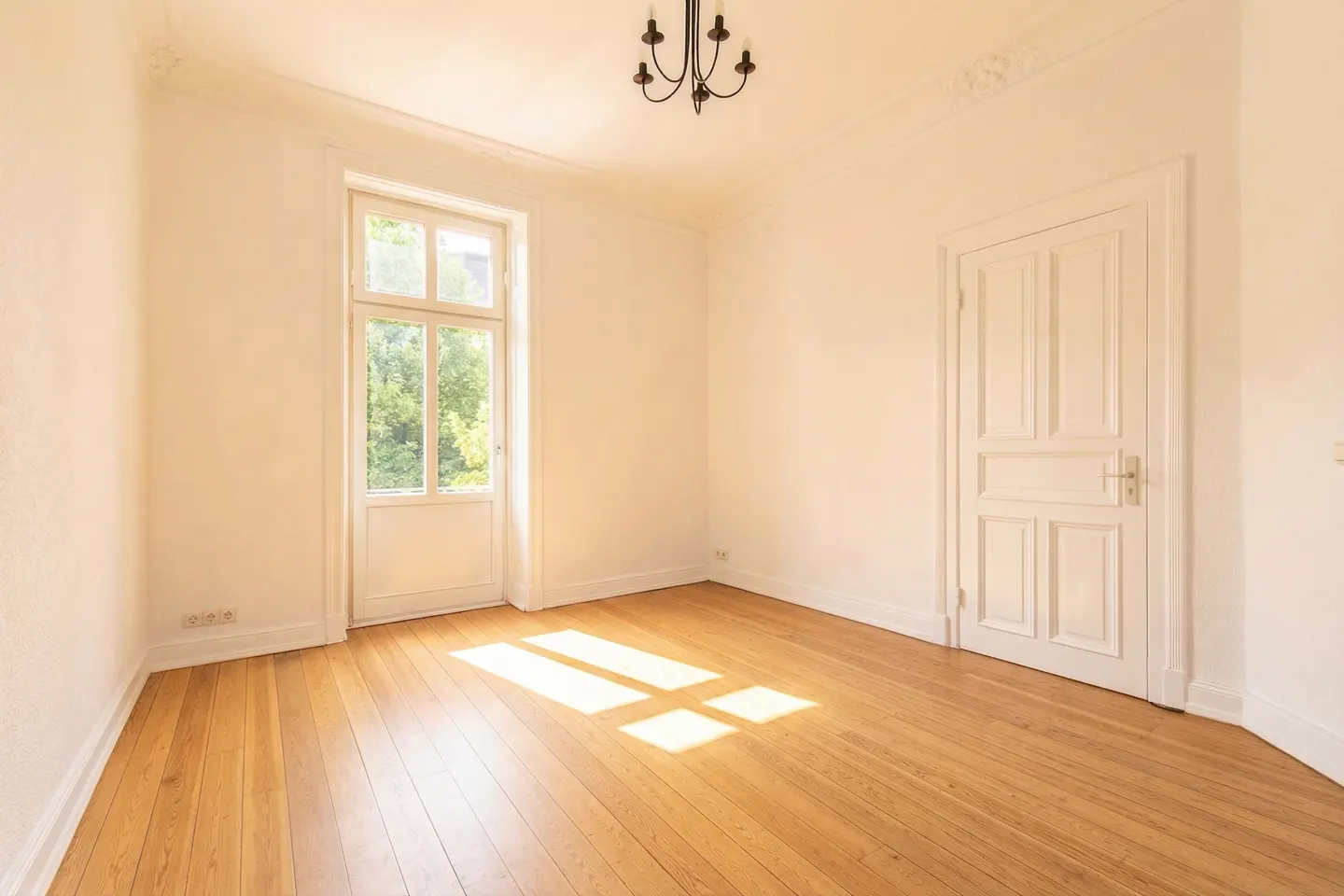 Bright, empty room with hardwood floors, white walls, and a window letting in sunlight. A chandelier hangs from the ceiling.