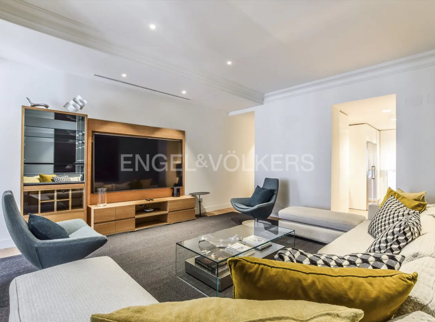 Bright living room with a large TV, modern furniture, and a glass coffee table. The room has a gray rug and white walls.