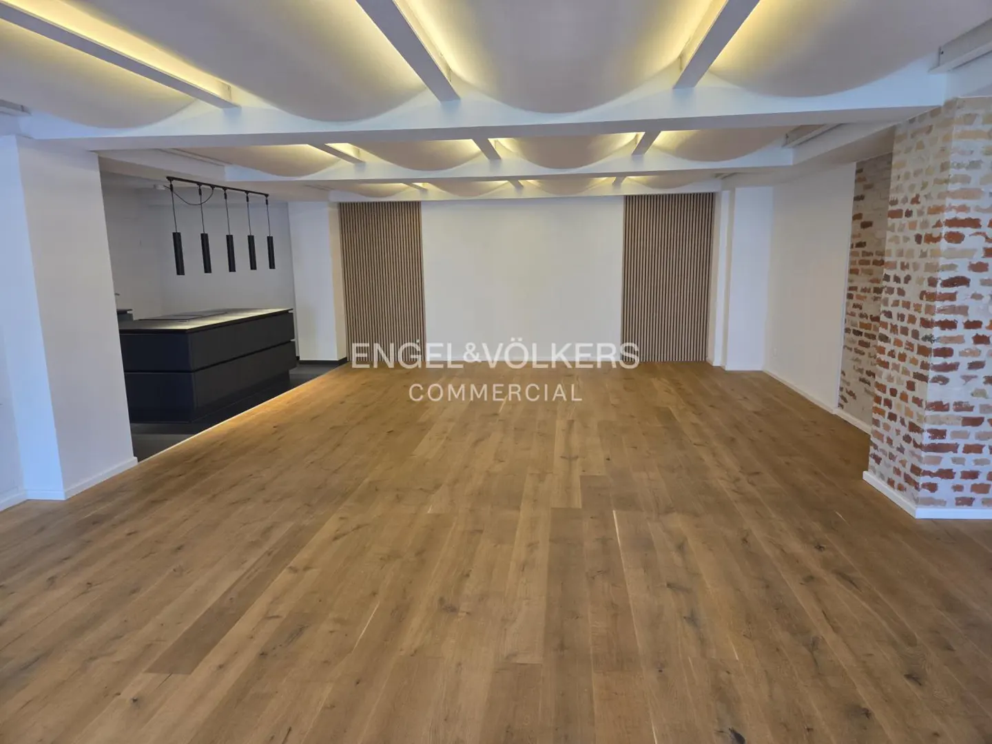 Bright, empty commercial space with wood floors, white walls, and exposed brick. Modern lighting and wood slat accents.