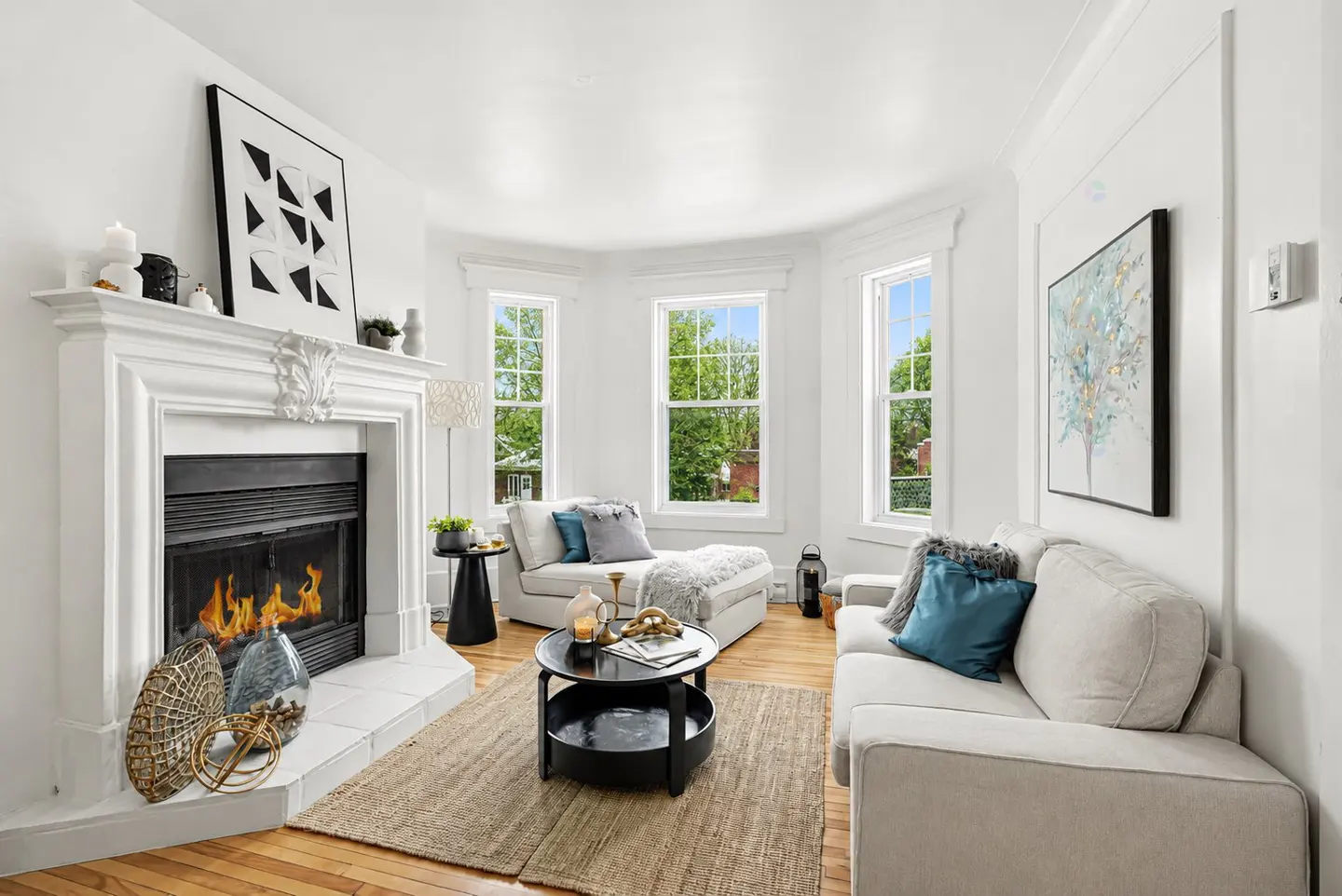 Bright living room with white walls, hardwood floors, and a fireplace with burning logs. Two white sofas face a black coffee table. Three windows let in natural light.