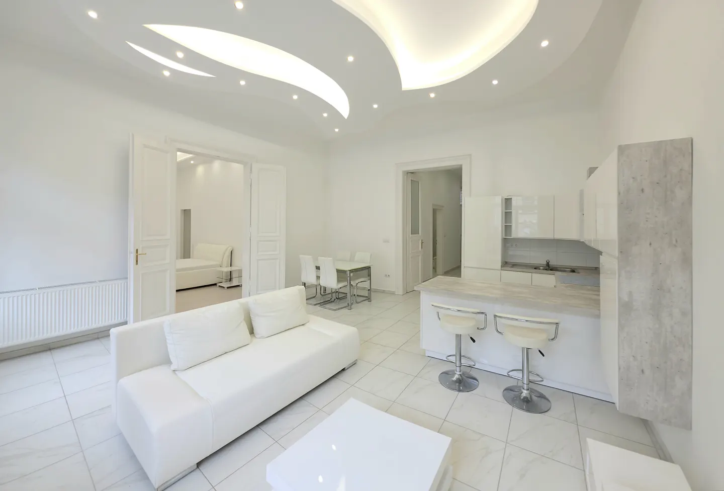Modern white open-plan living area with a sofa, dining set, kitchen island with bar stools, and a unique curved ceiling.