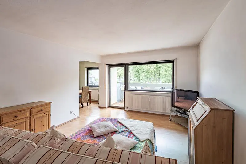 Bright living room with wood floors, white walls, and a striped couch. A colorful quilt and pillows are on the floor. A balcony door lets in natural light.