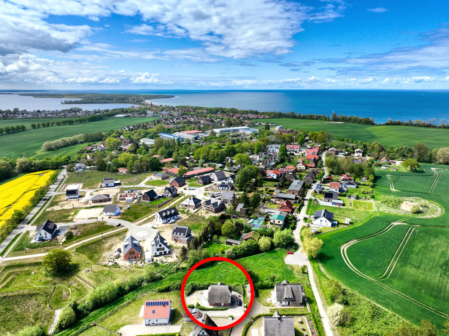Aerial view of a village with a thatched-roof house circled in red, near green fields and blue water under a partly cloudy sky.