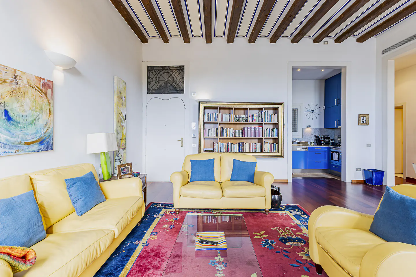 Bright living room with yellow sofas, blue pillows, and a red rug. A bookshelf and a blue kitchen are visible in the background.