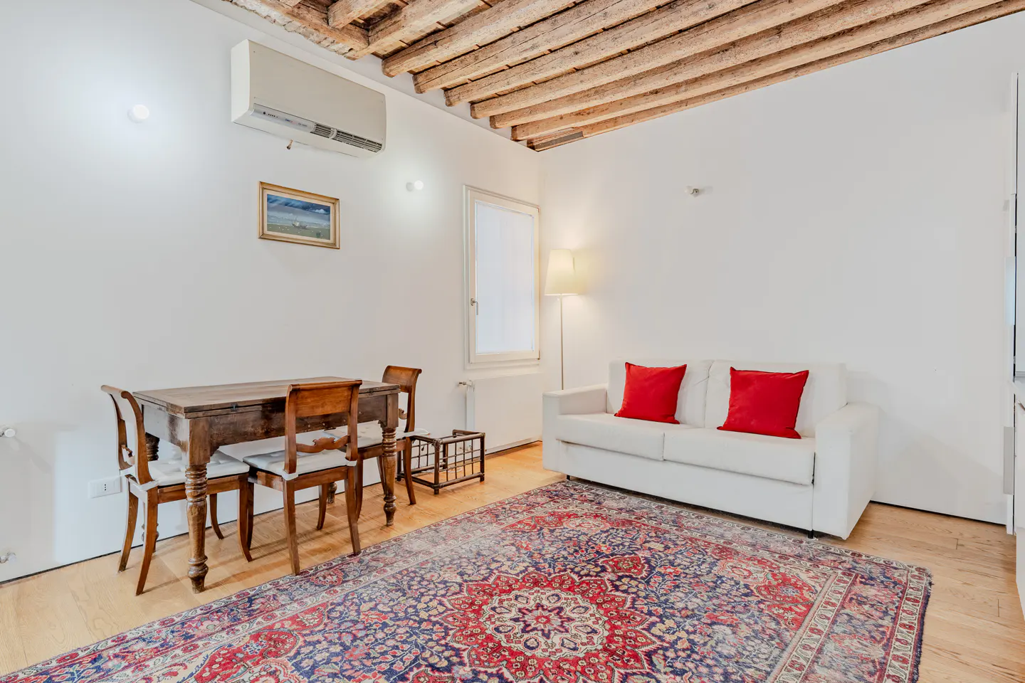 Living room with white sofa, red pillows, wooden dining table, and a large patterned rug under a rustic beamed ceiling.