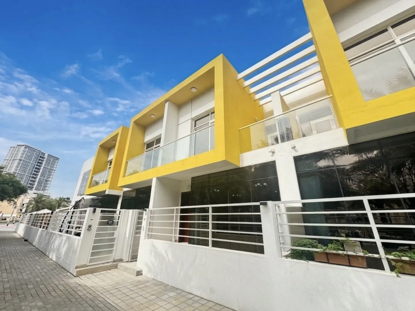 Exterior view of modern townhouses with yellow accents, white fences, and glass balconies under a blue sky.