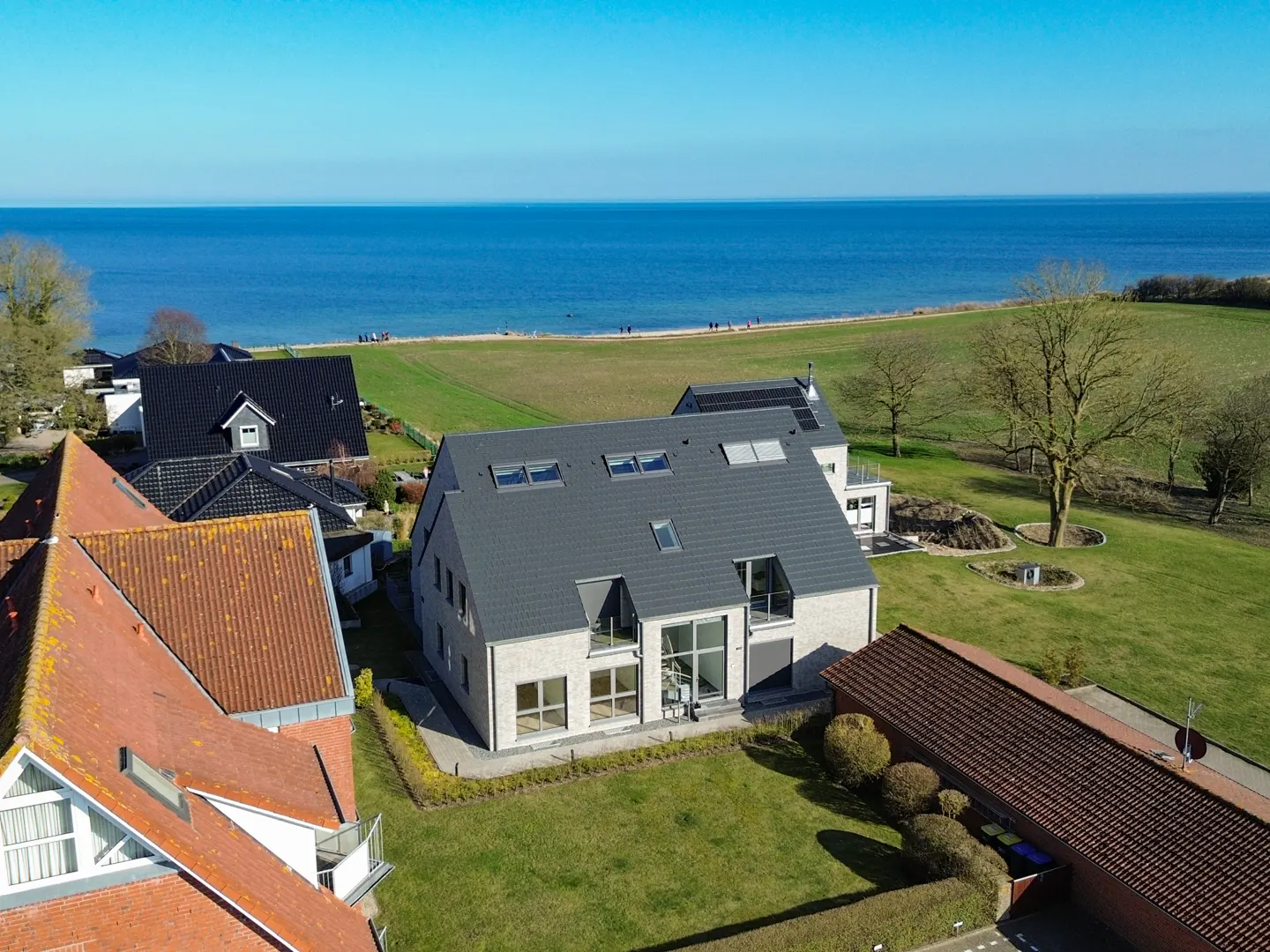 Aerial view of a modern, light-brick house with a gray roof, surrounded by green lawns and other houses, with the blue sea in the background.