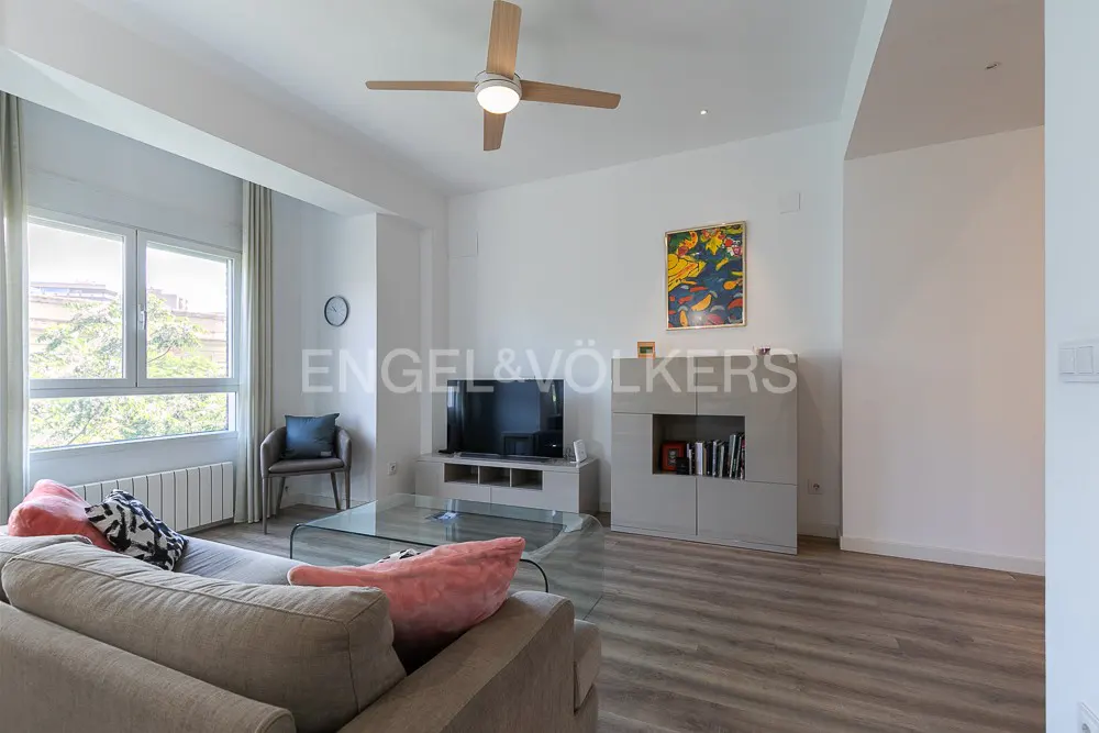 Bright living room with a gray sofa, glass table, TV, and a ceiling fan. A large window lets in natural light.