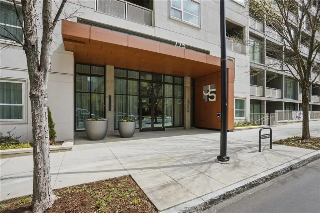 Exterior shot of a modern apartment building with a wood-paneled entrance and large windows. Two silver planters flank the entrance.