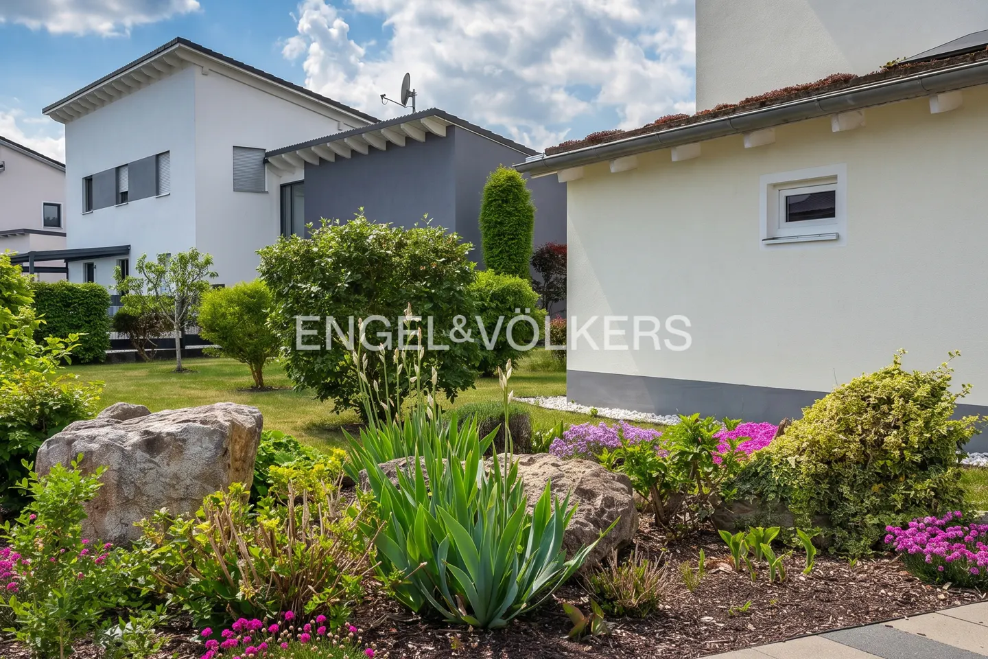 Landscaped yard with rocks, flowers, and bushes in front of modern white and gray homes under a blue sky. Engel & Völkers logo visible.