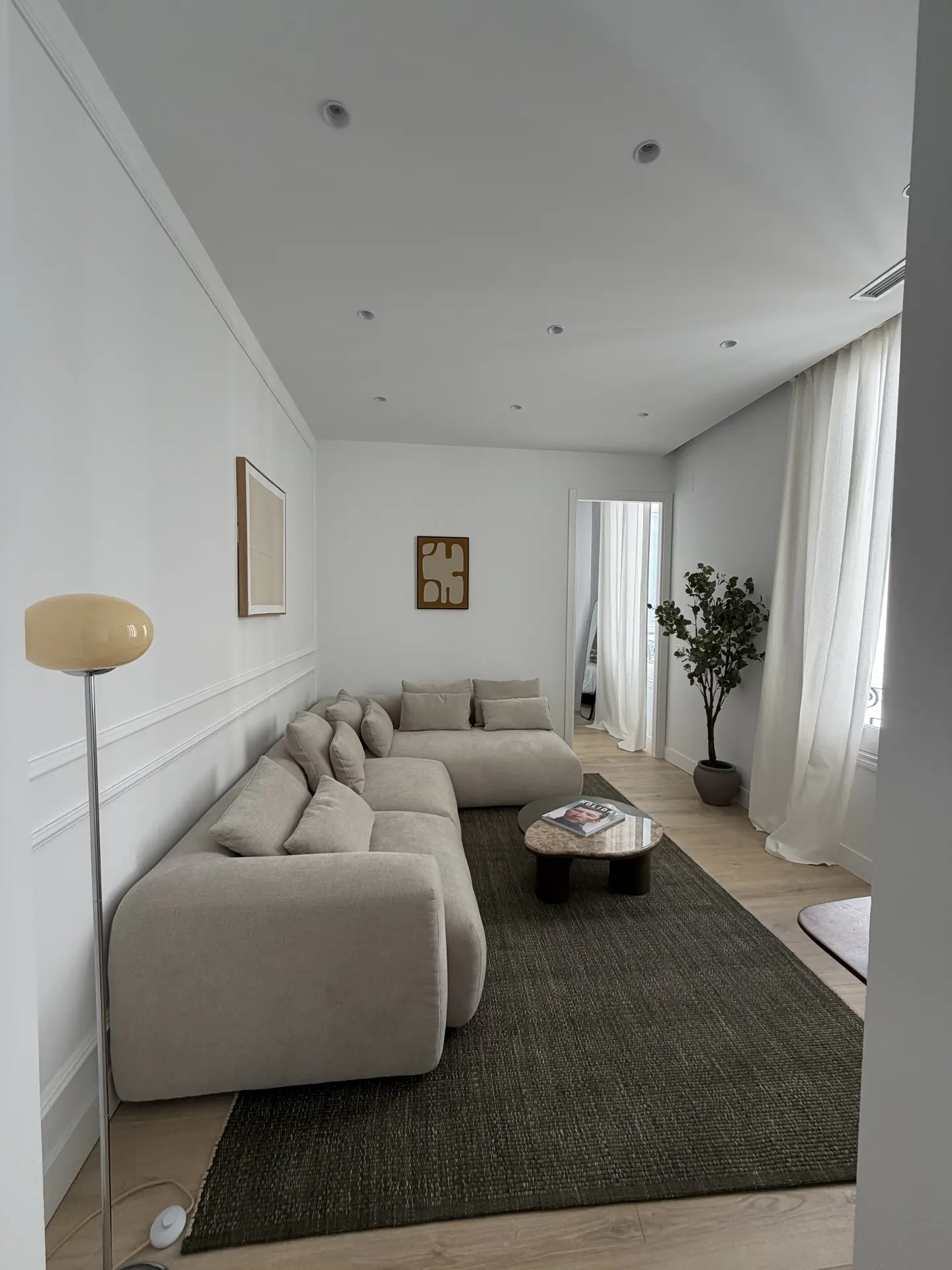 A bright living room with a large beige sectional sofa, a dark rug, and white walls. A floor lamp and coffee table are also visible.