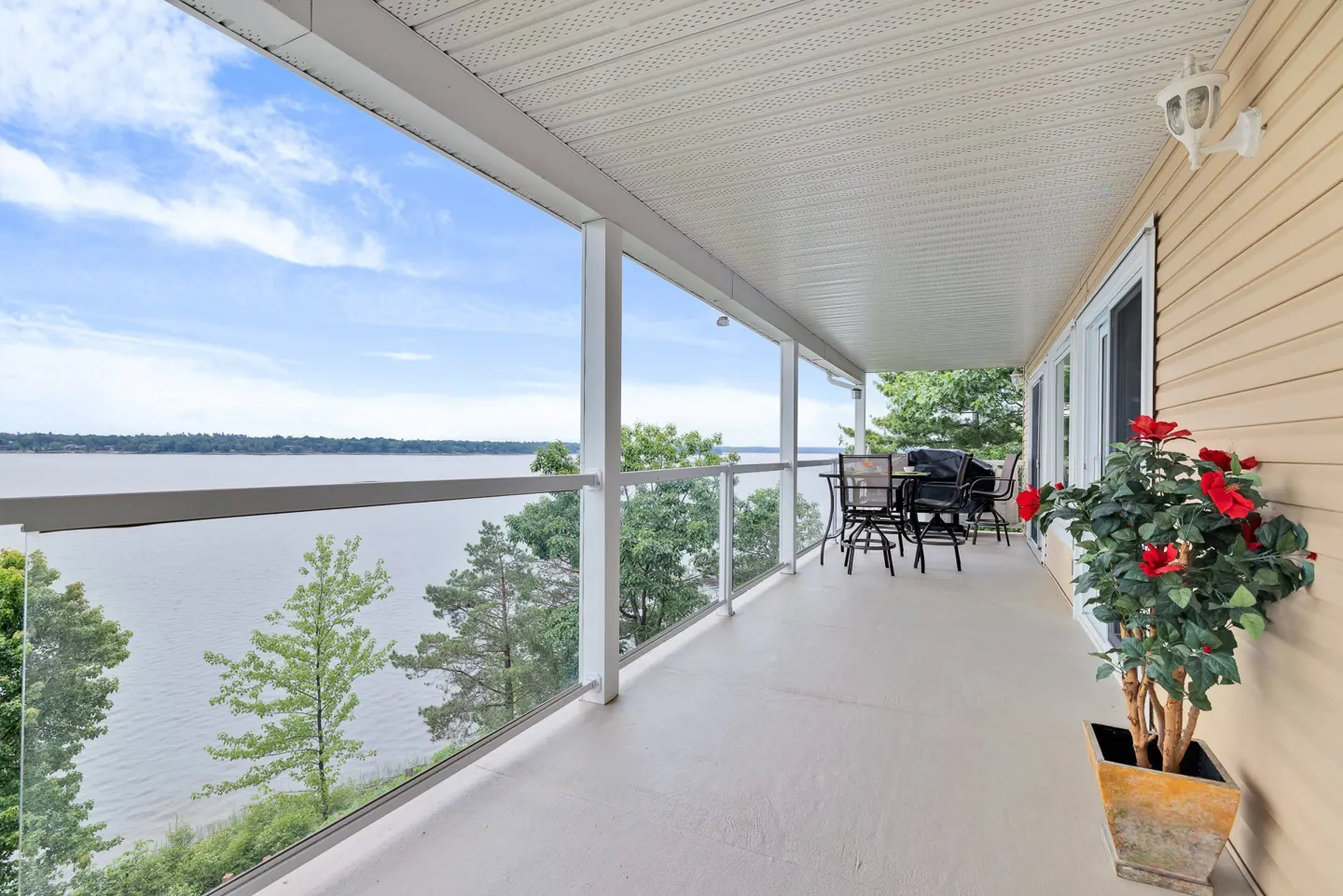 Covered balcony with glass railing, patio furniture, and a potted plant overlooking a scenic lake and trees.