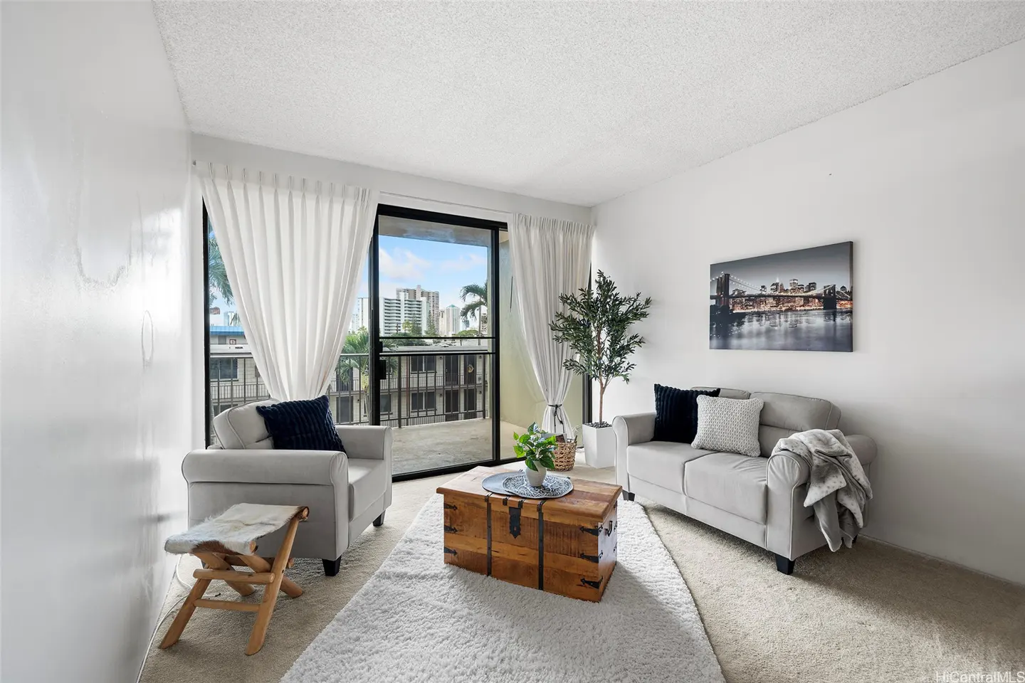 Bright living room with white walls, a gray sofa and chair, and a wooden chest on a white rug. Balcony with city view.