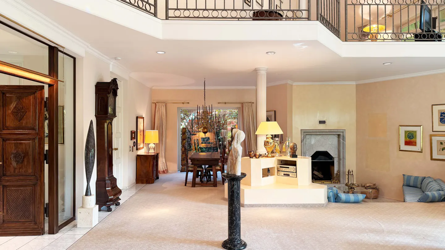 A spacious living room with a fireplace, dining table, and a second-floor balcony. The room is decorated with sculptures, lamps, and artwork.