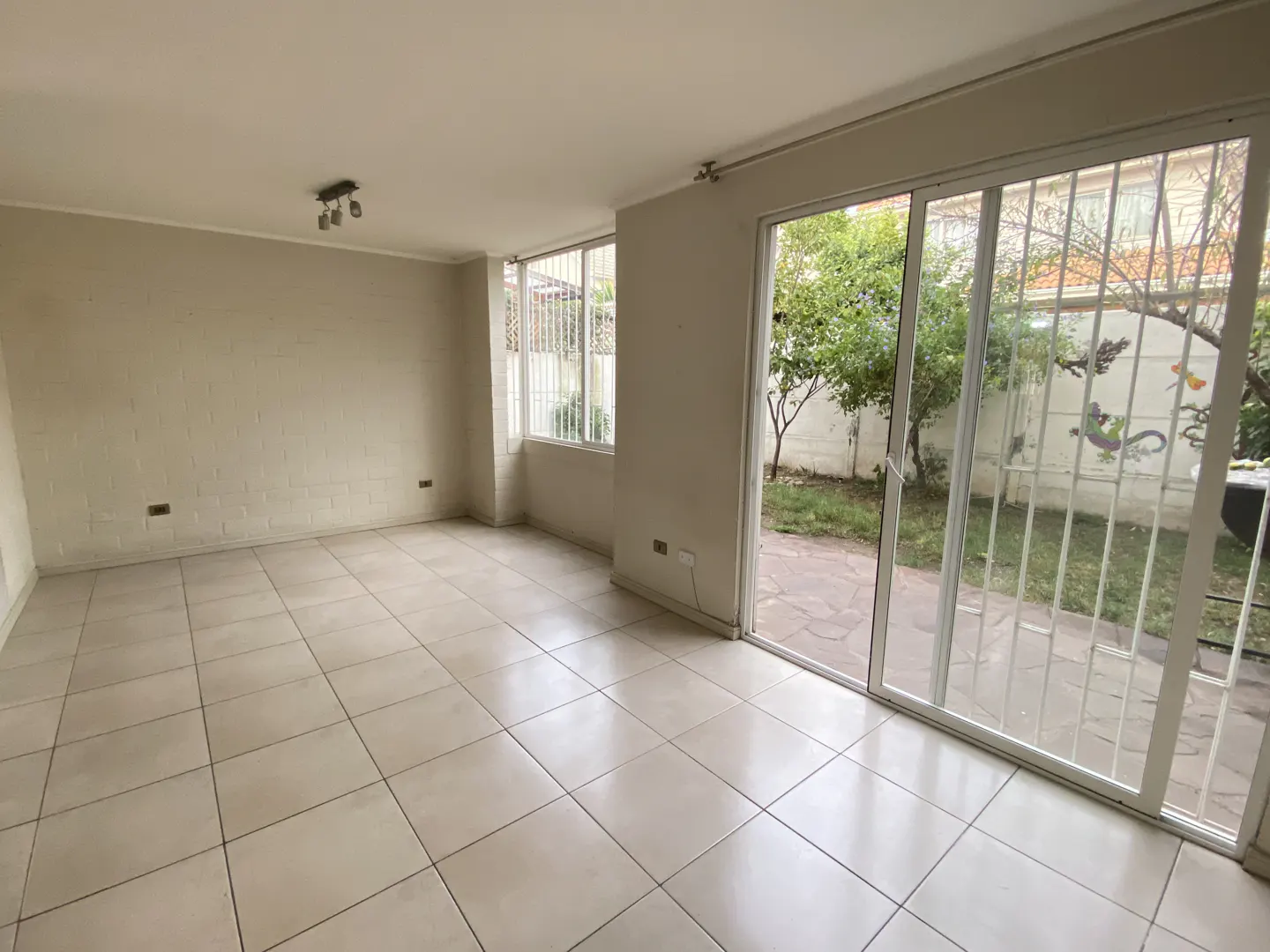 Empty room with beige walls and tile floor. Sliding glass doors open to a small yard with trees and a white fence.