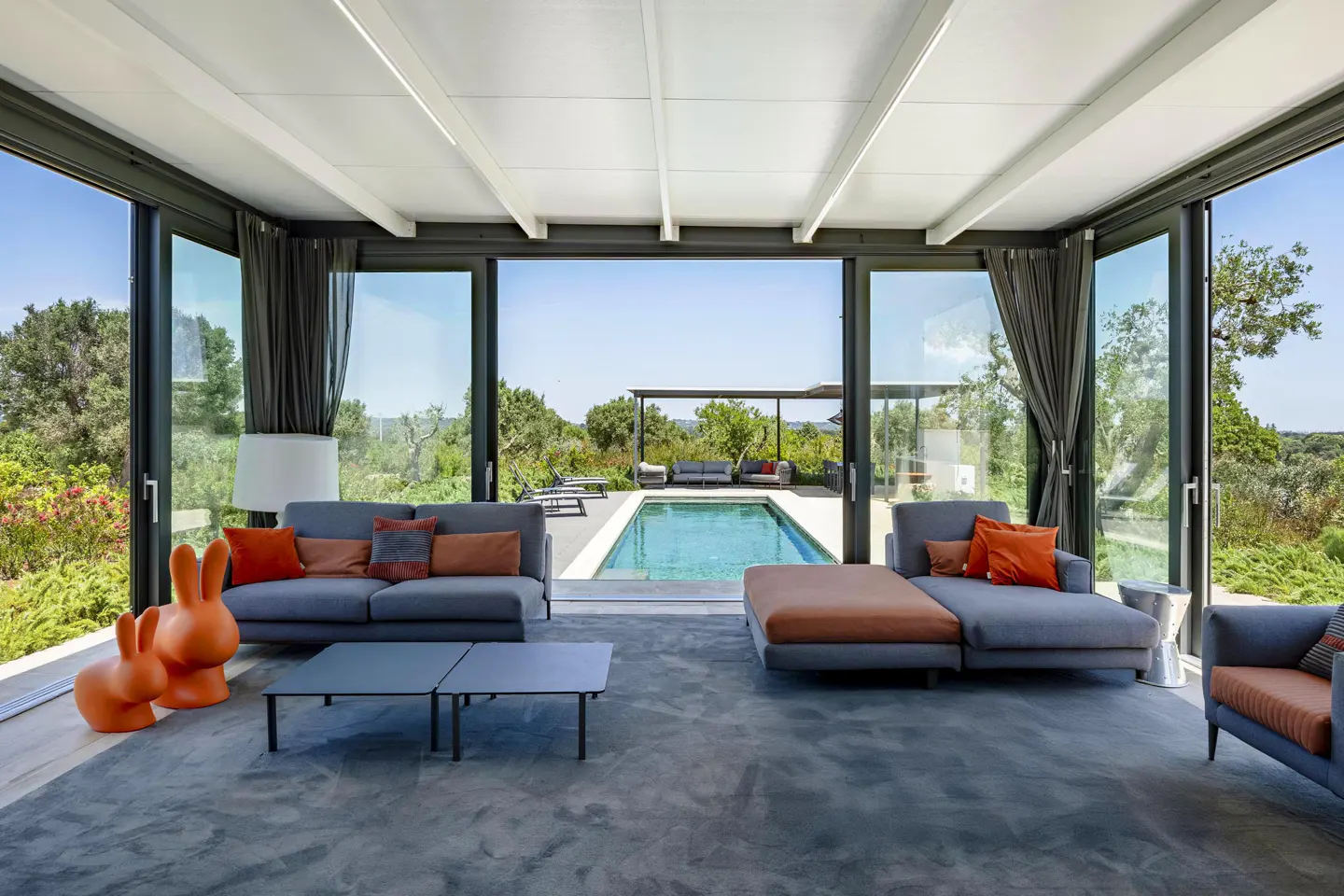 Modern living room with grey sofas, orange accents, and floor-to-ceiling windows overlooking a swimming pool and garden.