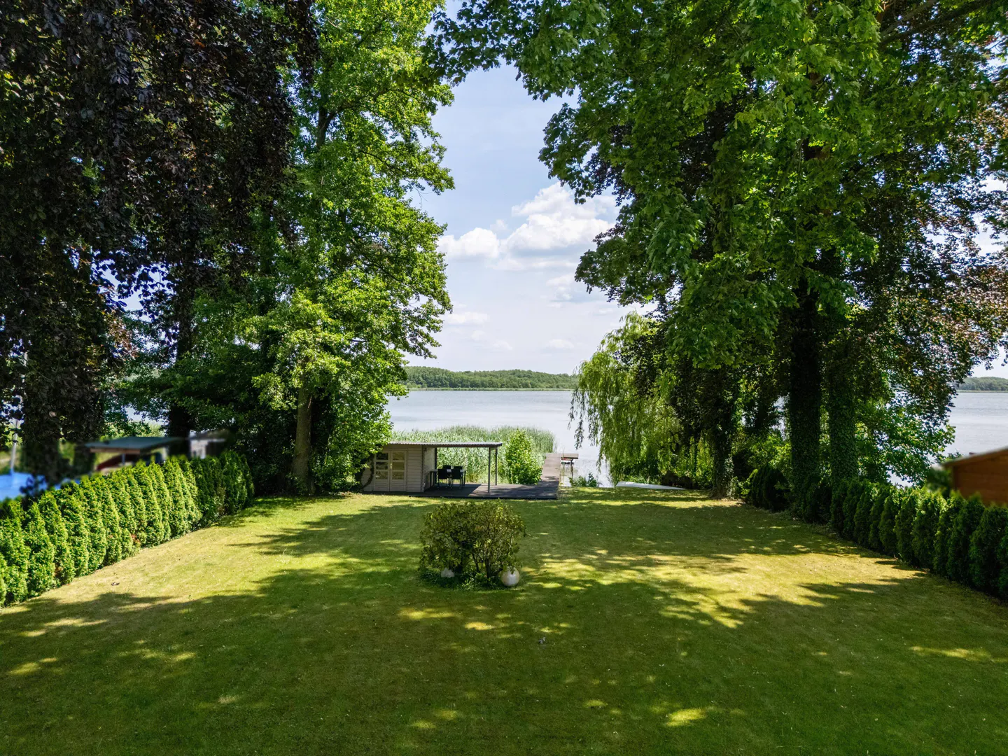 Lush green lawn with trees framing a lake view. A small dock and boathouse are visible on the water's edge.