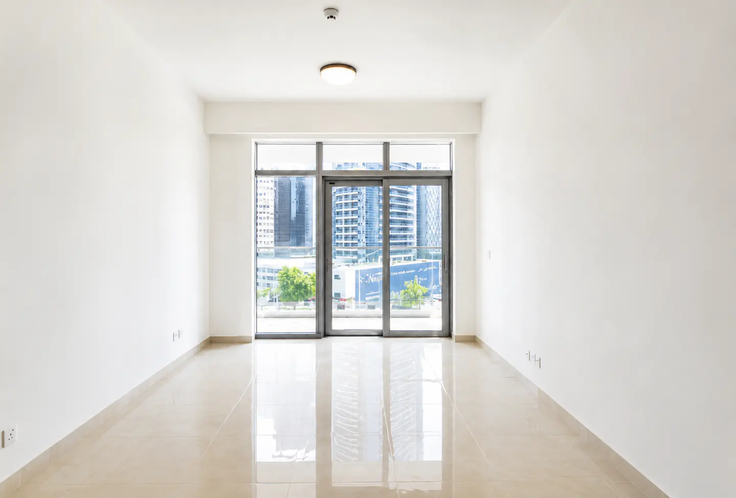Empty room with white walls and beige tile floor. Large window shows city buildings and trees. Ceiling light visible.