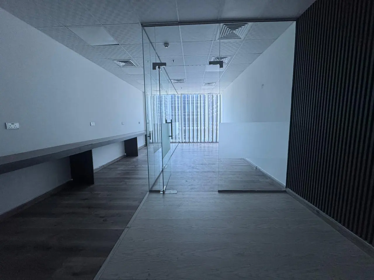 Empty office space with glass door, white walls, and wood floors. A long desk runs along one wall. Vertical blinds are visible through the glass.