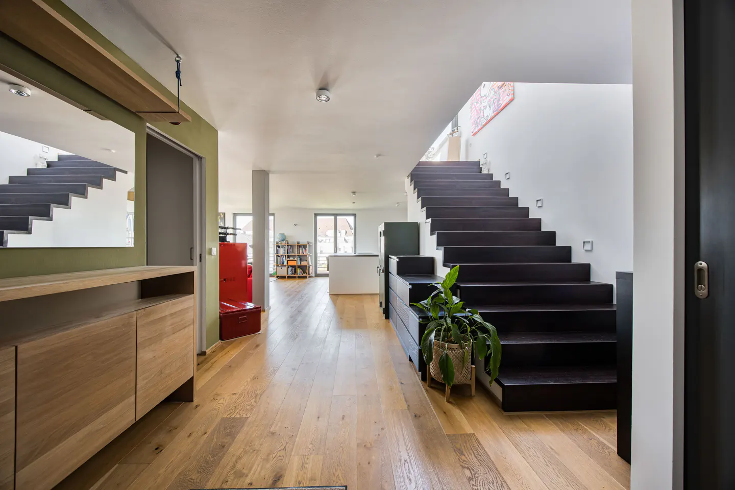 Modern home entryway with wood floors, dark stairs, and a light wood cabinet. A red refrigerator is visible in the background.