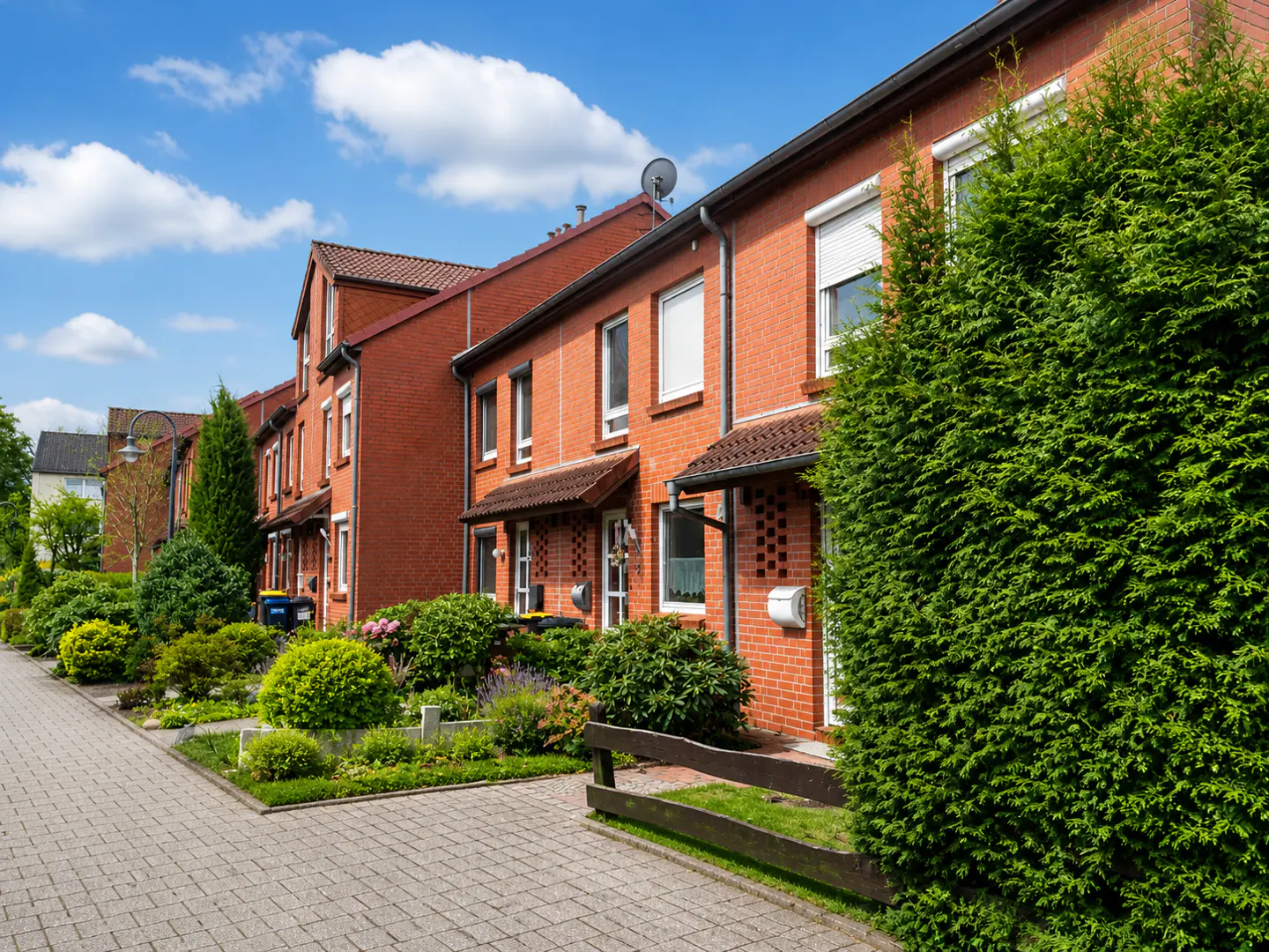 Row of red brick townhouses with green bushes and trees under a blue, cloudy sky.
