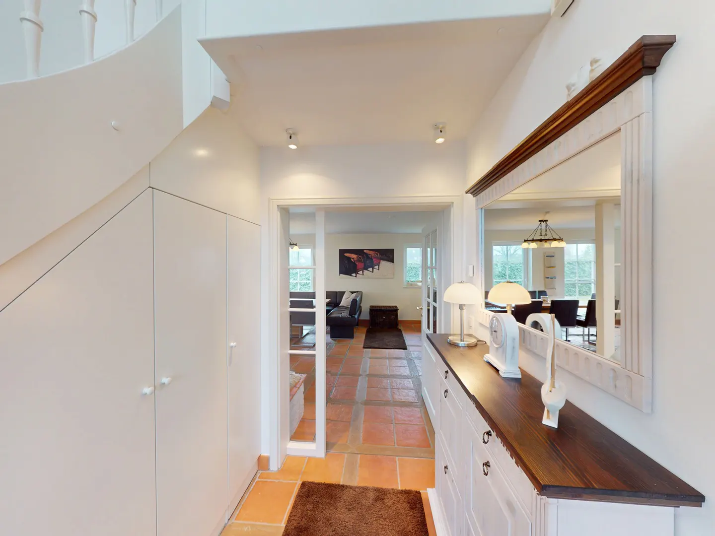 Hallway with white walls, terracotta tile floor, and white storage cabinets under a winding staircase. A mirror hangs above a dark wood-topped cabinet.