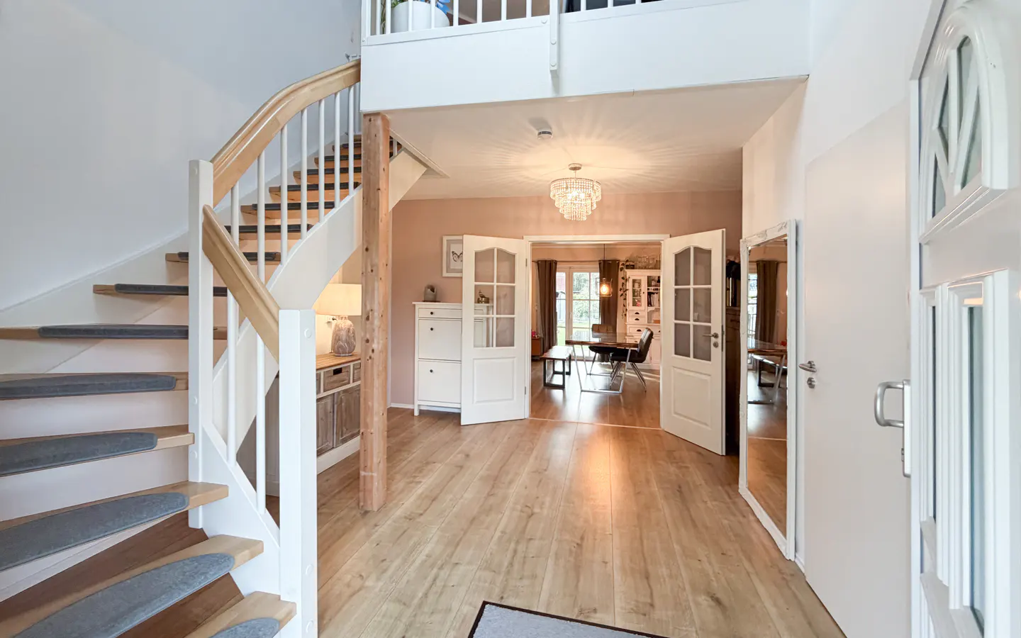 Bright foyer with wood floors, white staircase with gray treads, and open double doors to a dining room with a chandelier.